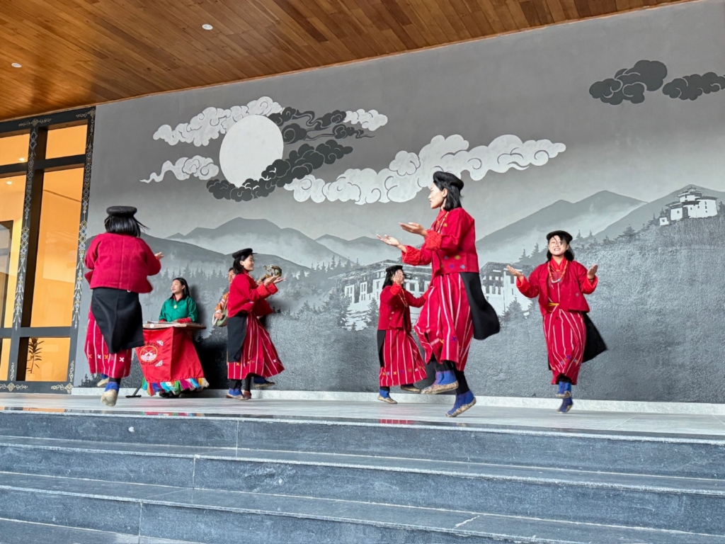 A group of Bhutanese women in matching red garments performing a graceful folk dance with synchronized hand gestures.