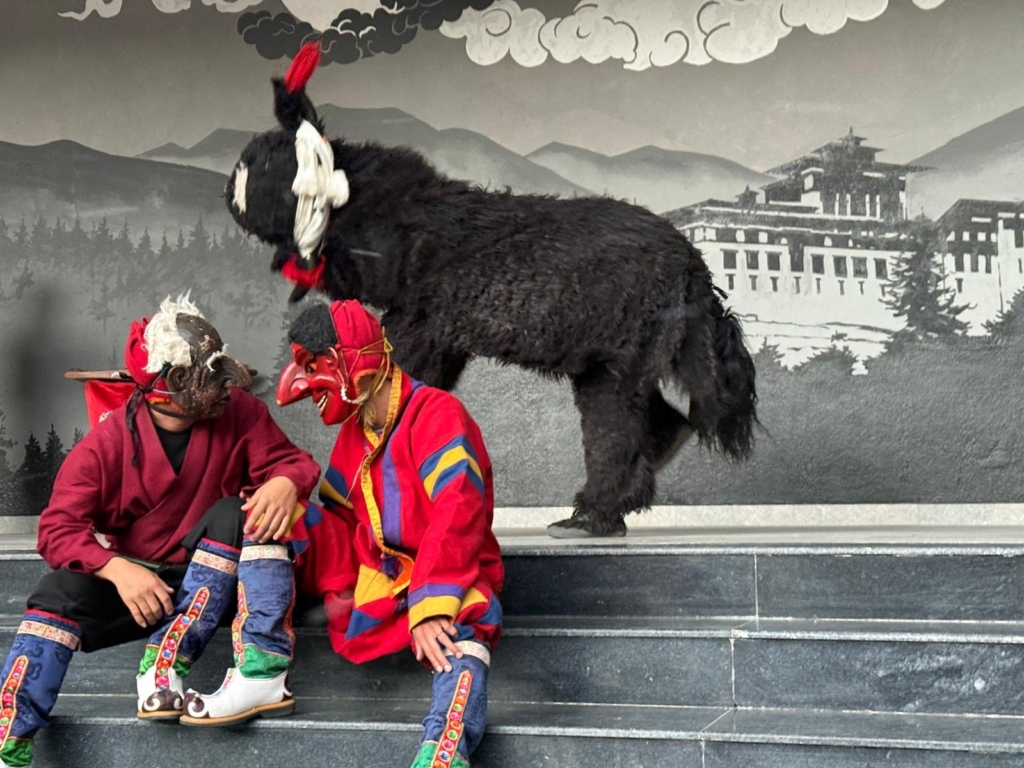 Two masked performers sitting casually on the stage steps, accompanied by a large, shaggy black yak costume.