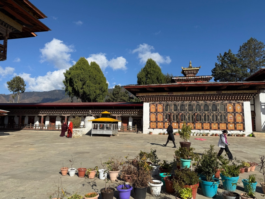 The spacious, sunny inner courtyard of Gangtey Goenpa featuring potted plants and a row of prayer wheels, while monks walk across the square.