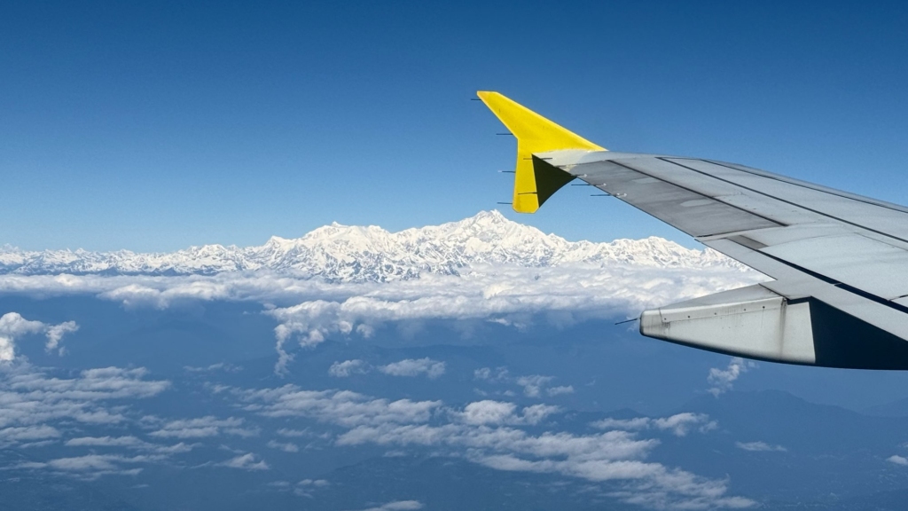 Blick aus dem Fenster eines Flugzeugs über eine dichte Wolkendecke auf die schneebedeckte Gebirgskette des Himalaya. Im Vordergrund ist die Tragfläche mit der charakteristischen gelben Spitze zu sehen.