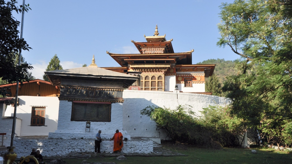 A monk in conversation in front of the white walls and dark roof of an outbuilding within the spacious grounds of the Punakha Dzong.