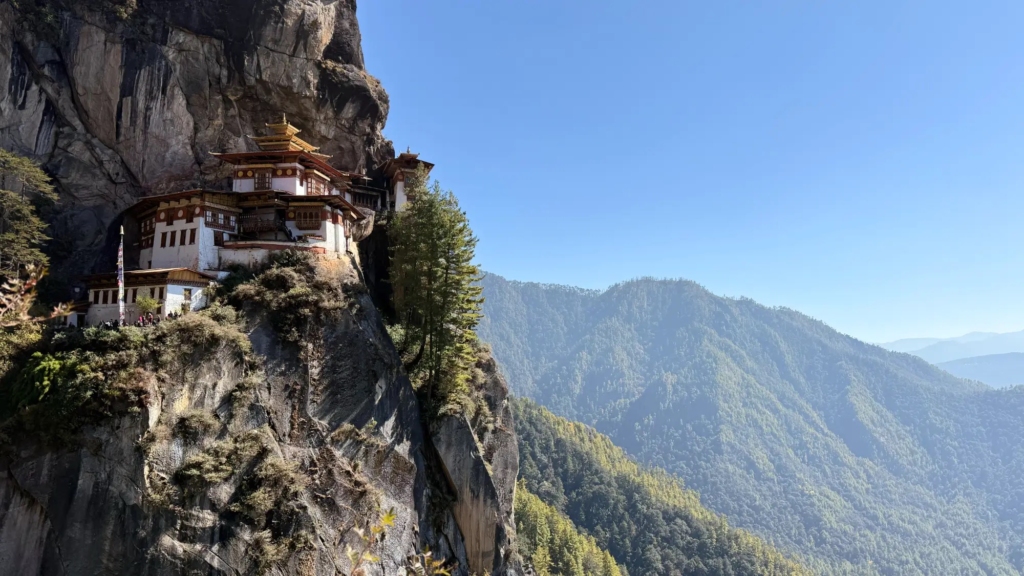 The famous Tiger's Nest Monastery (Paro Taktsang), clinging spectacularly to a sheer cliff face in the Paro Valley of Bhutan, set against a blue mountain backdrop.