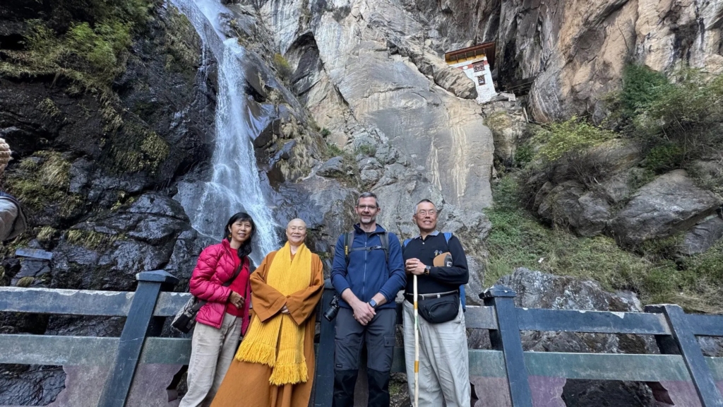 Gongjue Tuji and Venerable Master Shi Zheng Da standing with Dharma siblings in front of the sacred waterfall near the entrance to Paro Taktsang (Tiger's Nest).