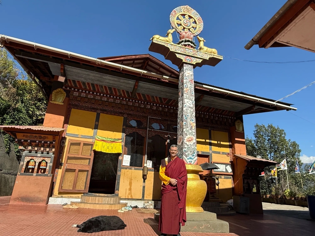 Gongjue Tuji in traditional robes standing in the sunny courtyard of the Thangton Dewachen nunnery next to a tall Dharma pillar, while a dog sleeps peacefully in front.