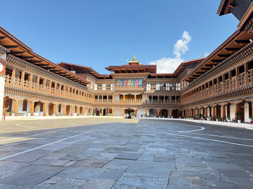 The spacious, paved inner courtyard of Wangdue Phodrang Dzong, surrounded by multi-story wooden galleries under a blue sky.